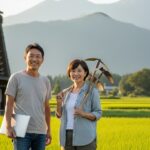 日本の田舎の集落で、地域おこし協力隊として活動する50代の男女が笑顔で立っている。後ろには茅葺き屋根の古民家と田園風景が広がり、ノートパソコンと農具を手に地域活性化に取り組む様子が描かれている。