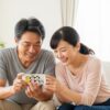 50代くらいの日本人の男女ペアが、明るいリビングのテーブルで向かい合って座り、一台のスマートフォンを二人で仲良く覗き込みながら、ディズニーツムツム風のゲームをプレイしている様子。二人は自然な笑顔を浮かべ、リラックスした雰囲気でゲームに夢中になっている。