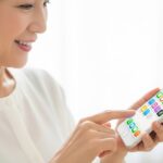 自宅のソファで複数のスマホゲームアプリのアイコンが並んだ画面を見ながら笑顔でスマートフォンを操作している50代の日本人女性の写真 。