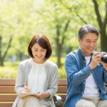 50代の日本人男女が公園で趣味を楽しんでいる様子。男性はカメラで写真を撮り、女性はベンチでスケッチを描いている。