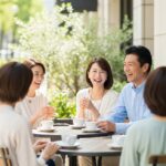 50代の日本人男女が屋外カフェで笑顔で会話をしている様子。穏やかな雰囲気の中で人間関係のつながりを感じるシーン。