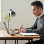50代の日本人男性が自宅のワークスペースでノートパソコンに向かい、キャリアについて考えながらノートを取っている様子。
