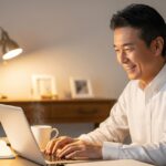 自宅の机に座り、ノートパソコンで質問に笑顔で答えている50代の日本人男性。親しみやすく思慮深い表情で、部屋は暖色系の照明で整頓されており、ノートとコーヒーカップが近くにある。読者からの質問に答えたり、アドバイスを提供したりしている様子。