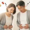中年の男女がお腹に手を当てて体型を気にしている様子。背景には野菜やダンベルなど中年太りに関連するアイテムが浮かび、健康意識を連想させる構成。
