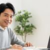 ノートパソコンでオンライン読書会に参加している50代の日本人男性が、明るい室内で本を読みながら微笑んでいる様子。画面には複数の参加者の顔が映り、和やかな雰囲気。
