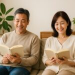 50代の日本人夫婦が明るいリビングで笑顔で読書を楽しんでいる様子。落ち着いた雰囲気の中で、前向きな気持ちと自己成長を感じさせる一枚。