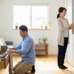 明るいリビングで、中年夫婦が収納の整理整頓をしている様子。夫は引き出しに毛布をしまい、妻は本棚に本を並べている。