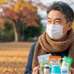 秋の公園でマスクとマフラーを着けた中年男性が、ビタミン剤やお茶、消毒液などの健康対策グッズを手に持っている。